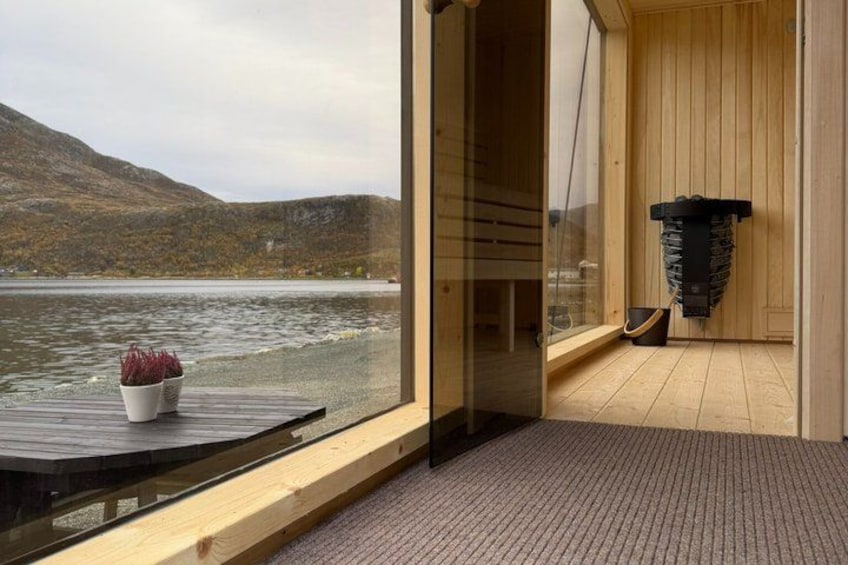A wooden cabin with a glass door leading to a sauna, overlooking a lake with mountains in the background. 3