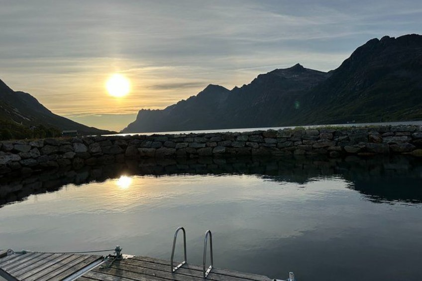 A calm lake with a wooden dock and stone wall, surrounded by mountains under a setting sun. 2