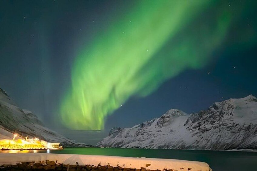 The Northern Lights over a snowy mountain range with a lit-up cabin in the foreground. 1