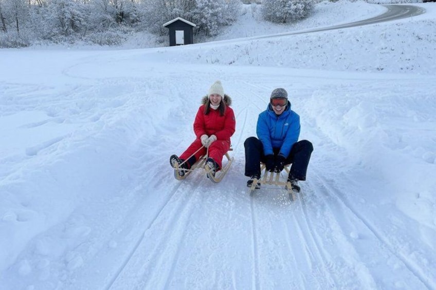 Two people sledding on a snowy slope with a small building in the background. 5