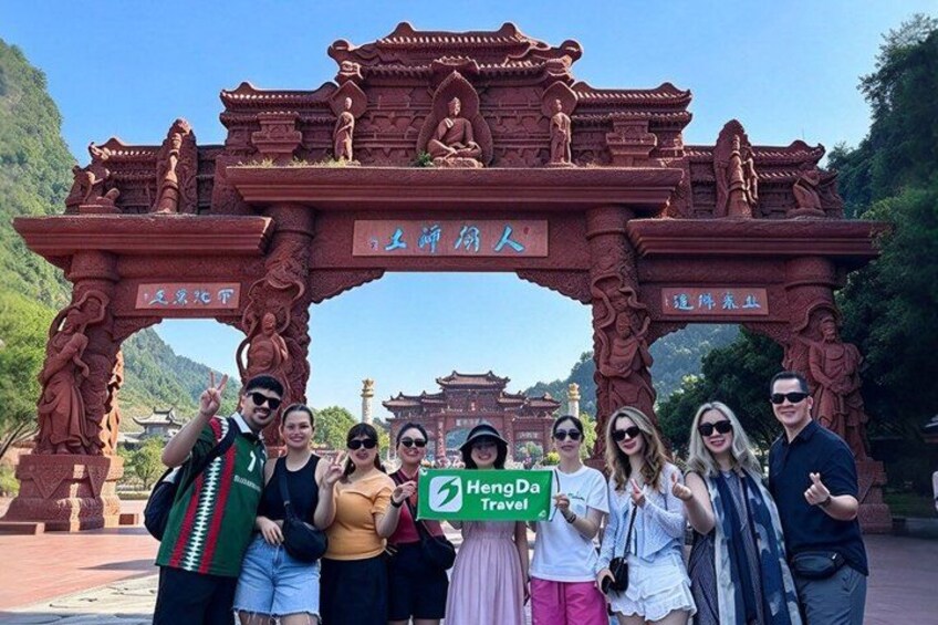 A group of people posing in front of a red archway with inscriptions, surrounded by greenery and a clear sky. 4