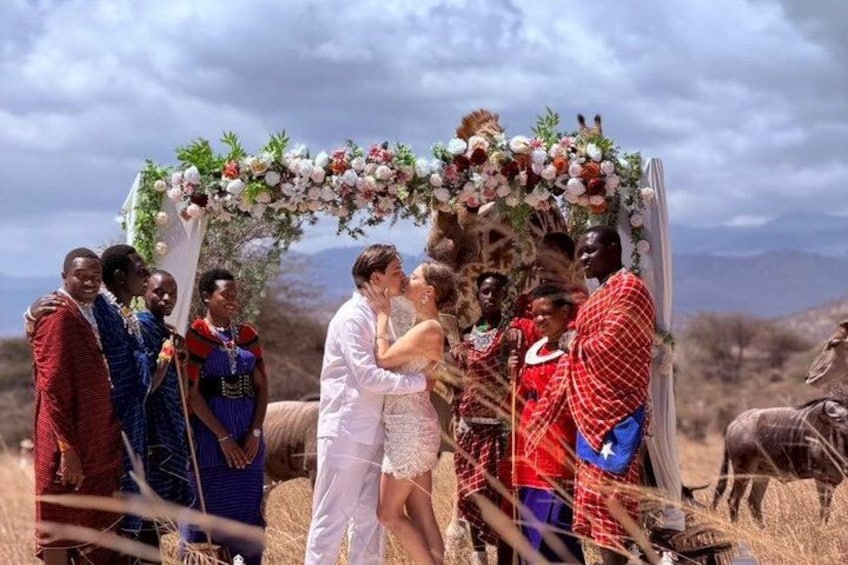 A traditional ceremony with a floral arch, people in traditional attire, and a backdrop of a dry landscape. 1