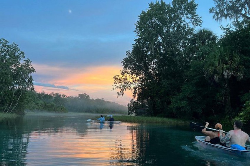 A calm river with people paddling kayaks, surrounded by dense trees and a sunset. 3