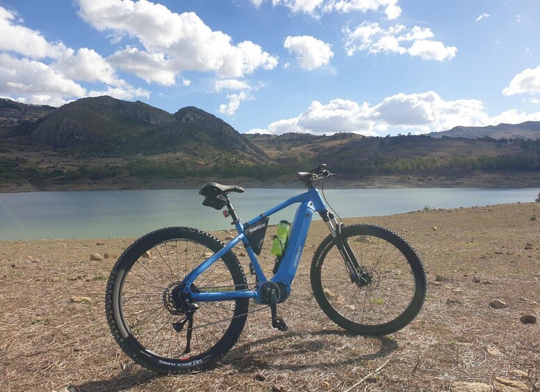 Una bicicleta de montaña azul con la marca Shimano en el cuadro, estacionada en un terreno seco y cubierto de hierba, con un lago y montañas al fondo. 1