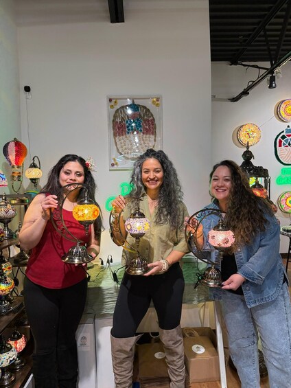 Three people holding decorative lamps in a store with various other lamps and a neon sign in the background. 1