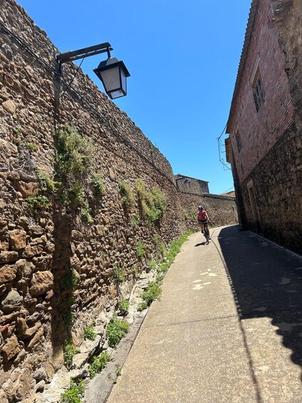 A narrow stone-paved street flanked by old stone walls, a cyclist in the distance, and a clear blue sky. 3