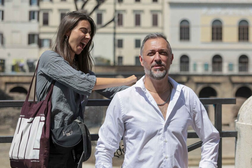 Un hombre con camisa blanca y una mujer con suéter gris de pie sobre un puente, con un edificio al fondo. 3