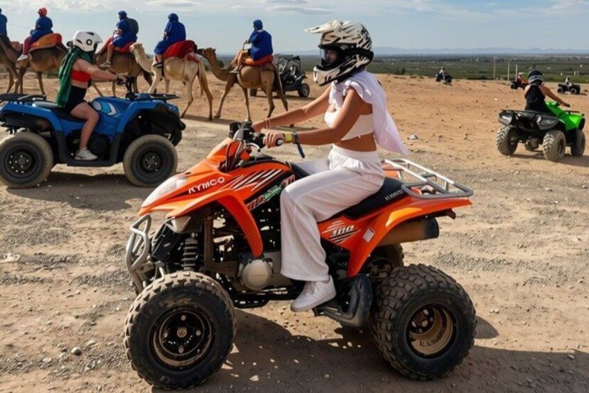 A person riding an orange ATV on a dirt road with camels and other ATVs in the background. 5