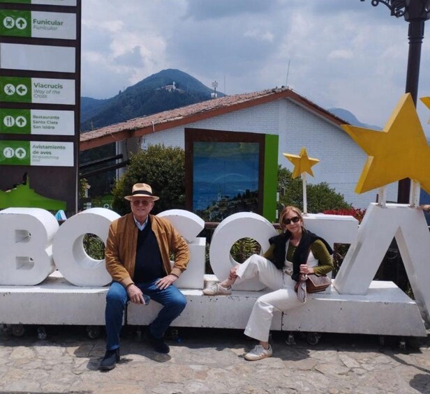 A couple sitting on a white structure with large letters spelling 'BOCA,' a mountain in the background, and a building with a red roof. 5