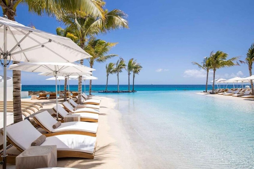 A beach resort with white lounge chairs and umbrellas, palm trees, and a clear blue ocean. 1