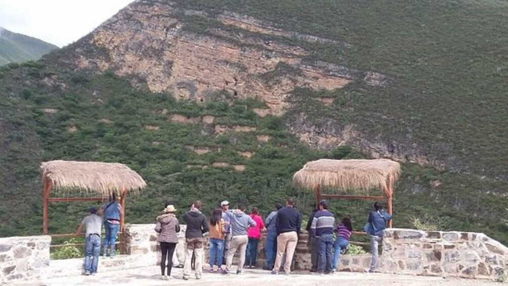 Un grupo de personas sobre una plataforma de piedra, con refugios de techo de paja, contemplando un paisaje montañoso. 5
