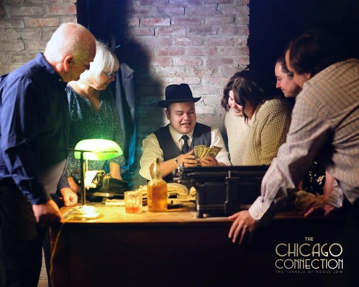 A group of people around a table with a lamp, a bottle, and a cash register. 1