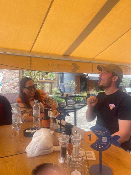 Two people seated at a wooden table with a yellow canopy overhead, a sign with a QR code, and a number 2 sign. 4