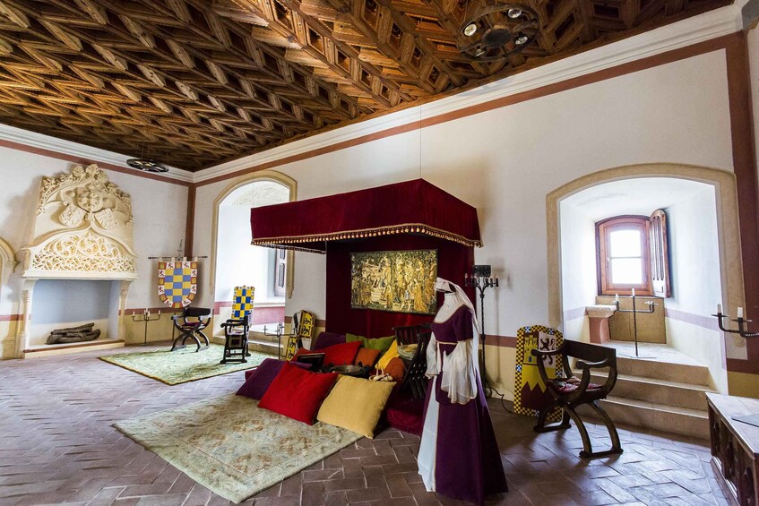 A medieval-style room with a wooden ceiling, a large bed with a red canopy, and a fireplace with a checkered cloth. 4