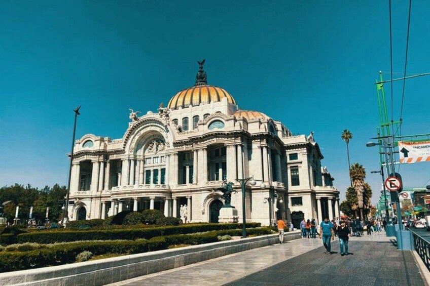 A large, ornate building with a golden dome and columns, surrounded by a well-maintained garden and a busy street with pedestrians. 1