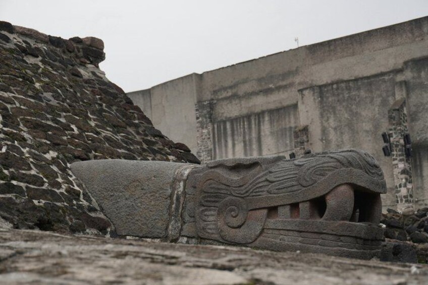 A stone carving with intricate designs, possibly of an animal, set against a backdrop of a large concrete wall and a pile of bricks. 5