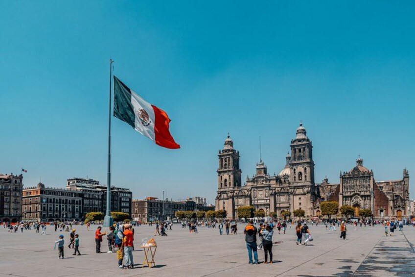 A large open plaza with a prominent flag, historic buildings, and a clear blue sky. 3
