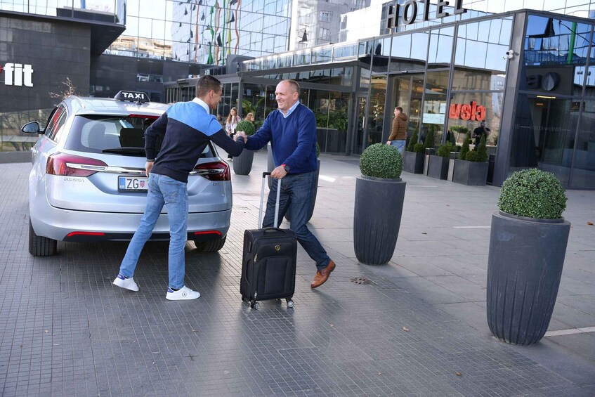 Two men shaking hands near a silver car with a 'TAXI' sign, in front of a hotel and a fitness center. 4