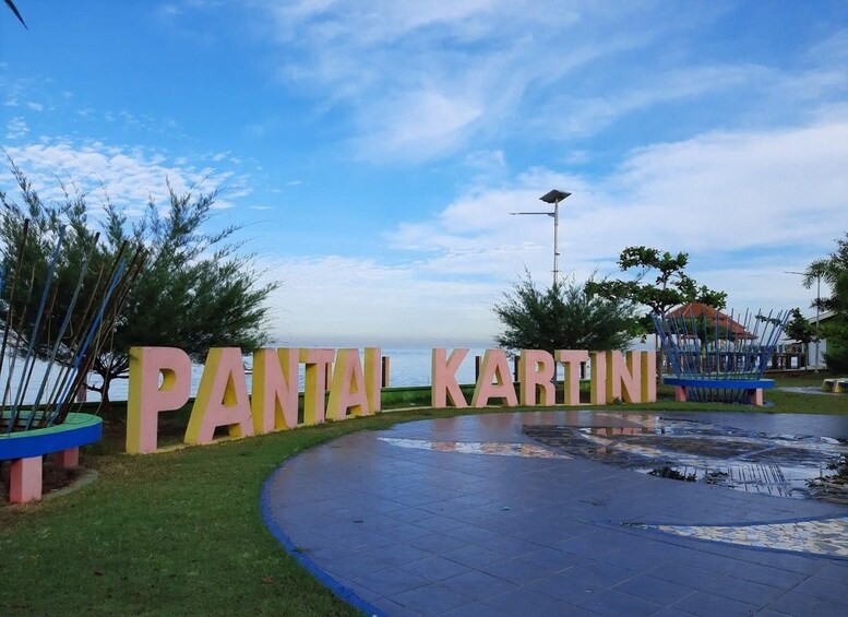 A sign with the words 'PANTAI KARTIN' in large letters, a paved walkway, and a body of water in the background. 2