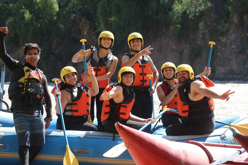 A group of people in a raft, wearing life jackets and helmets, on a body of water with trees in the background. 4