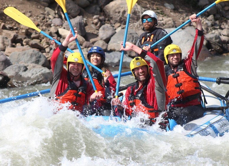 A group of people in a white raft with blue poles, wearing life jackets and helmets, navigating through white water rapids. 3