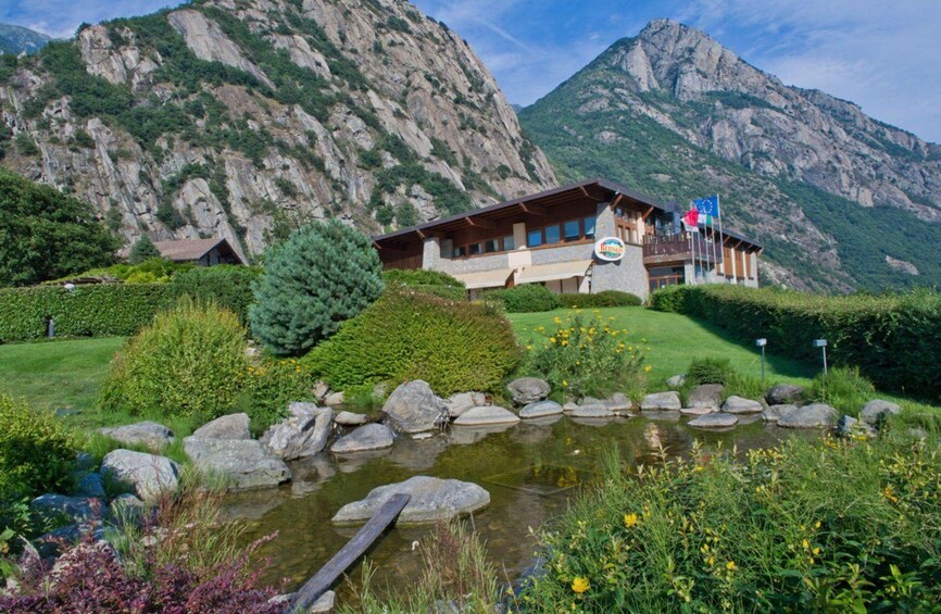 A mountain lodge with a pond, surrounded by lush greenery and rocky mountains. 2