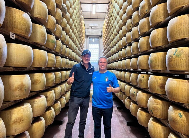 Two people standing between rows of large cheese wheels in a storage room. 3