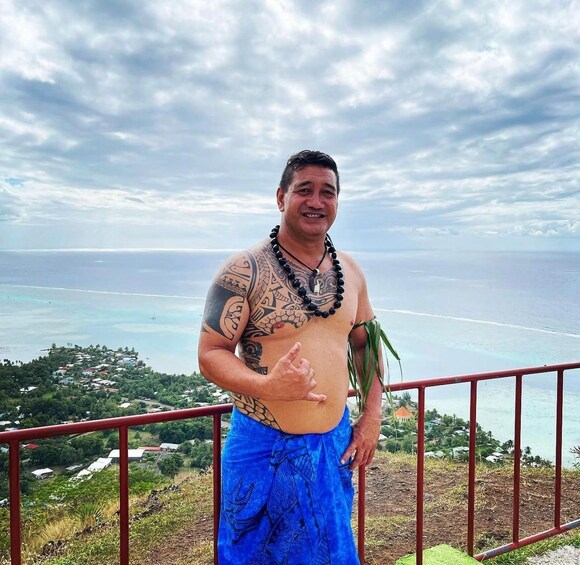 A man with tattoos standing on a balcony overlooking a coastal landscape. 2
