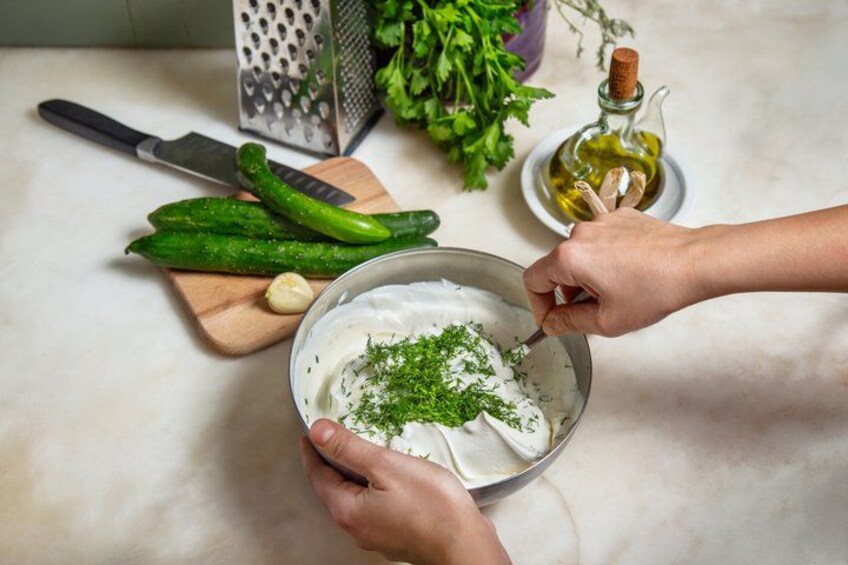 Hands mixing a creamy substance with herbs in a bowl, with cucumbers, garlic, and a grater on a cutting board, and a bottle of oil in the background. 5