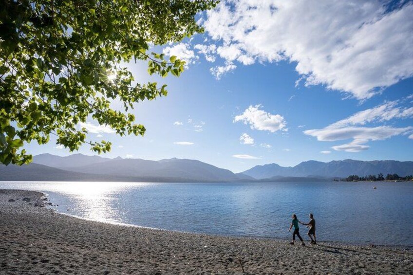 A lake with mountains in the background, a clear sky, and two people walking on the shore. 3