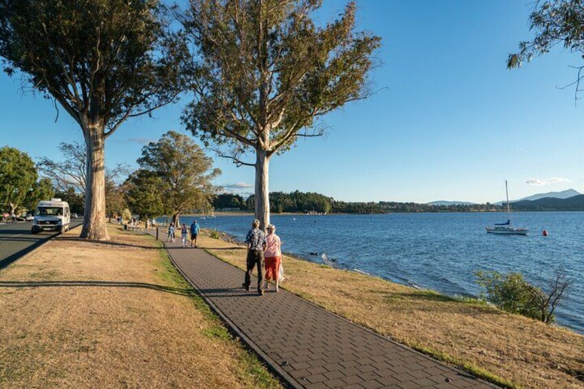 A lakeside path with people walking, trees, and a sailboat. 2
