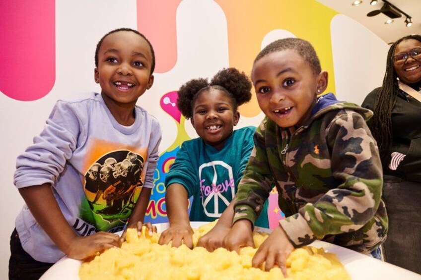 Three children at a table with a large container of yellow food, possibly mashed potatoes, in a brightly colored room with a playful background. 4