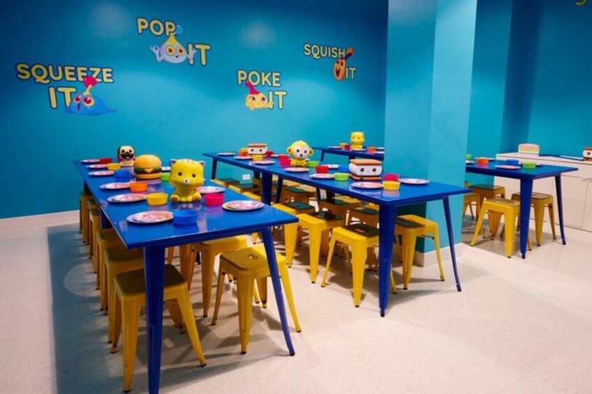 A brightly colored room with blue tables and yellow chairs, decorated with cartoon characters and the words 'POP,' 'SQUEEZE IT,' and 'POKE IT.' 2