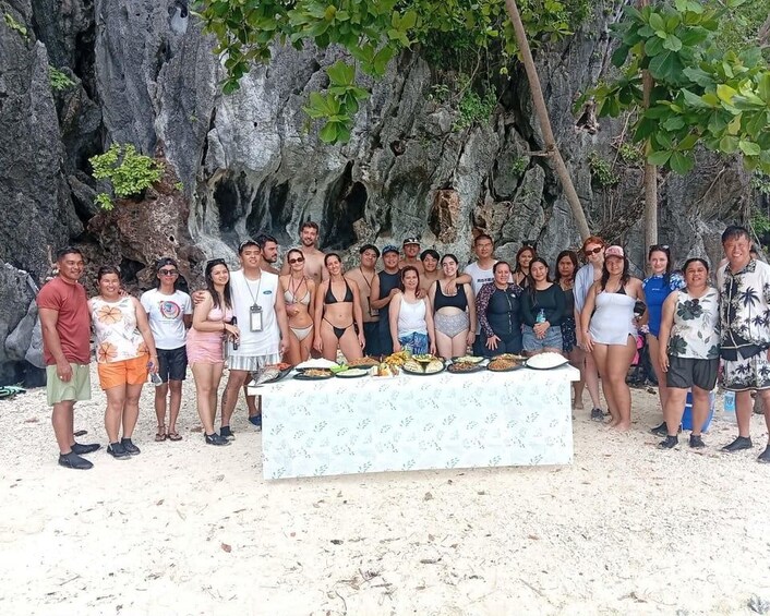 A group of people posing in front of a table with food on a beach with rocky cliffs in the background. 2