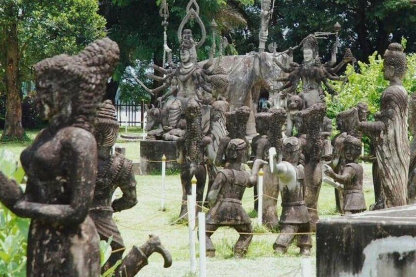 A stone statue of a deity surrounded by smaller figures, set in a garden with trees and a fence. 5