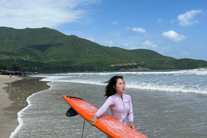 A person holding a surfboard on a beach with waves and green hills in the background. 2