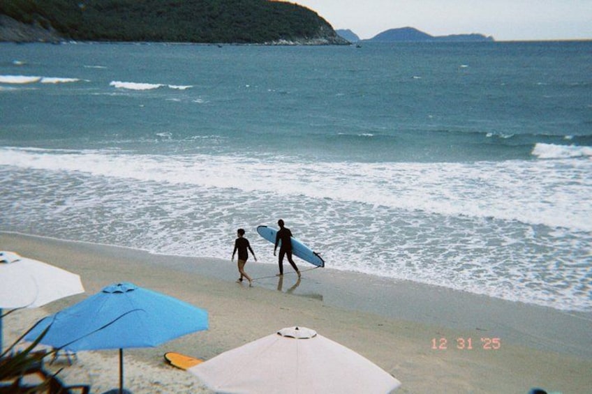 A beach with two people walking with surfboards, umbrellas, and a date stamp. 4