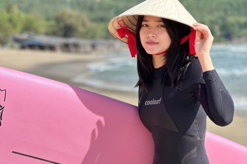 A person wearing a wetsuit and a conical hat, holding a pink surfboard, standing on a beach with the ocean in the background. 3