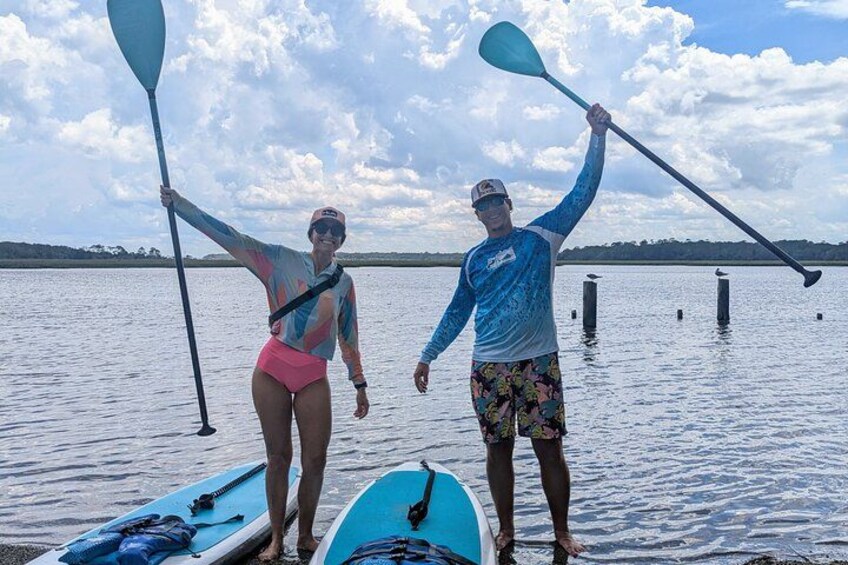 Two people standing on a dock with paddleboards and paddles, a calm lake, and a cloudy sky. 5