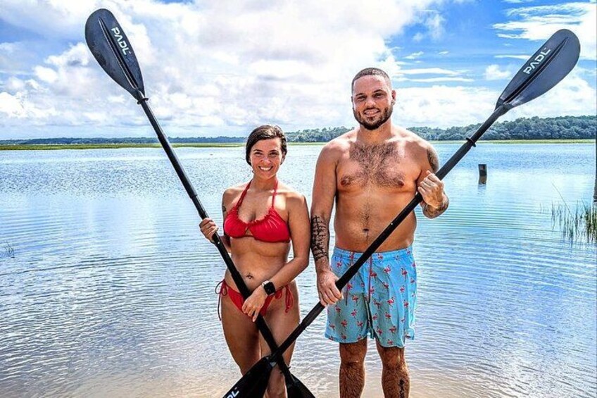 Two people holding paddles, standing by a calm lake with a clear sky. 1