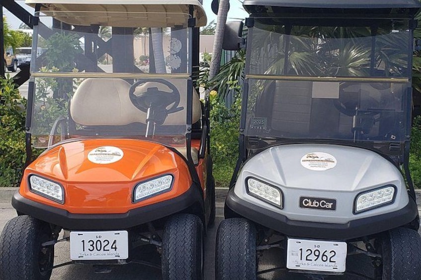 Two golf carts parked side by side, one orange and one gray, with license plates and Club Car branding. 2