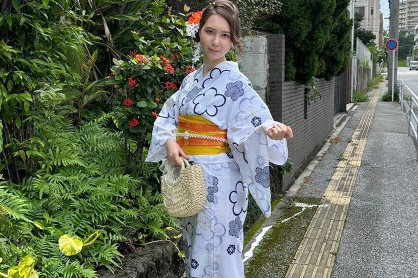 A person in traditional Japanese attire standing on a sidewalk with greenery and a brick wall in the background. 1