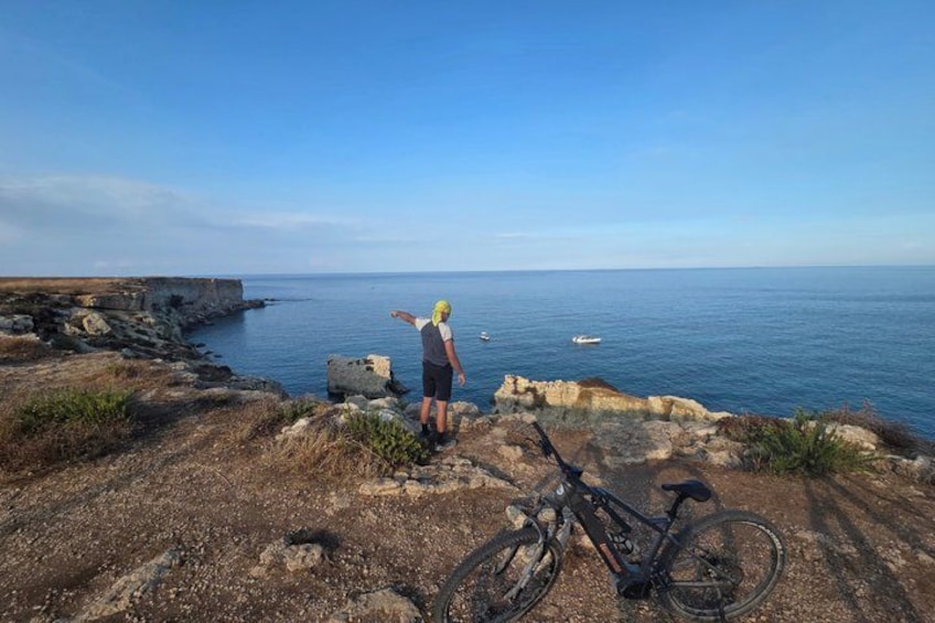 A person standing on a rocky cliff overlooking the ocean, with a bicycle parked nearby. 1