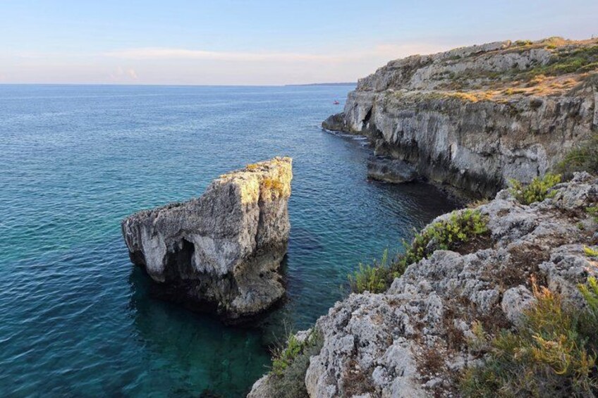 A coastal landscape with a prominent rock formation in the sea, clear blue water, and a rocky cliff. 3