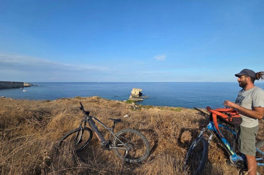 A person standing next to a bicycle on a grassy cliff overlooking the sea. 5