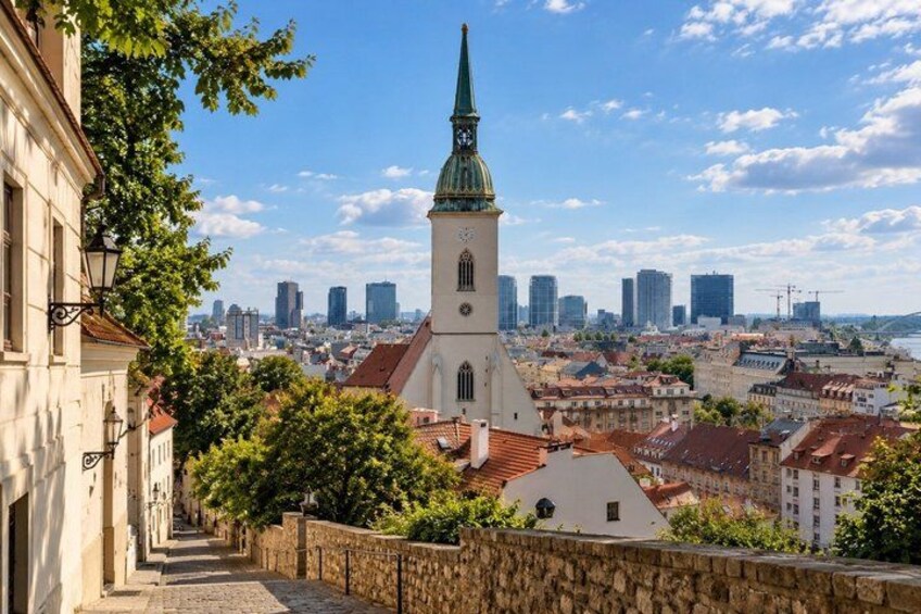 A historic church with a tall spire, a stone wall, and a cityscape with modern buildings in the background. 2