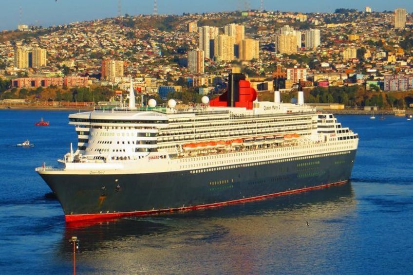 A large cruise ship named 'Queen Mary 2' docked near a city with a densely populated hillside in the background. 1