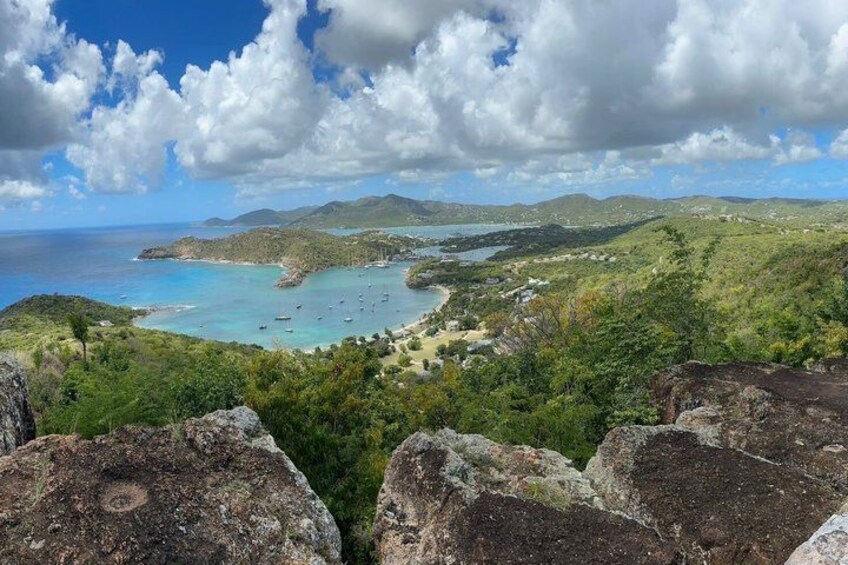 A coastal landscape with rocky outcrops, a bay with clear turquoise waters, and distant hills. 1