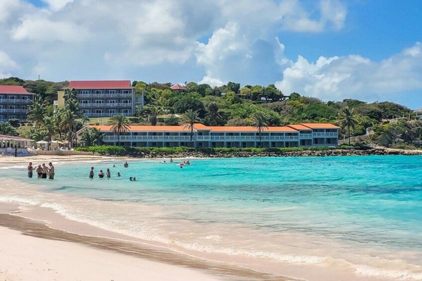 A beach with clear turquoise water, people swimming, and a hotel with a red roof in the background. 2