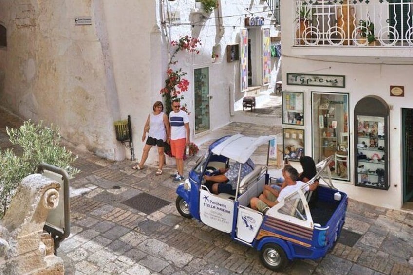 A narrow street with a blue and white car, pedestrians, and a shop with a sign. 1